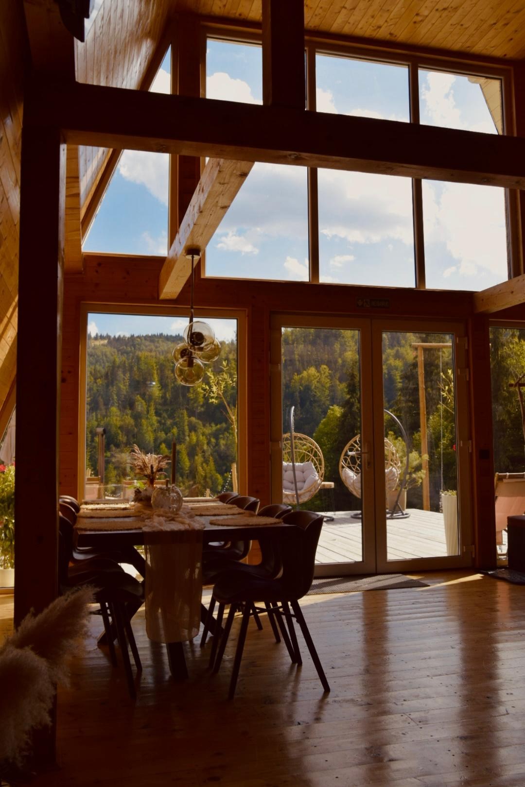 Masa din zona de dining, cu ferestre înalte spre terasă și pădure
